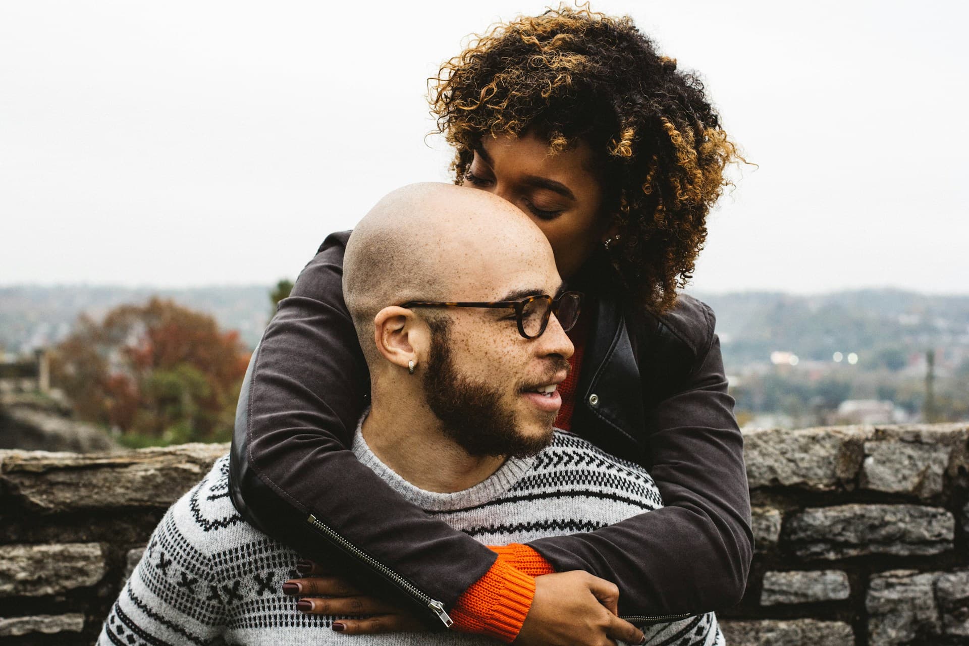 A couple embracing affectionately, with one partner gently kissing the other’s head. They are outdoors on a cloudy day, creating a warm and intimate moment of connection.