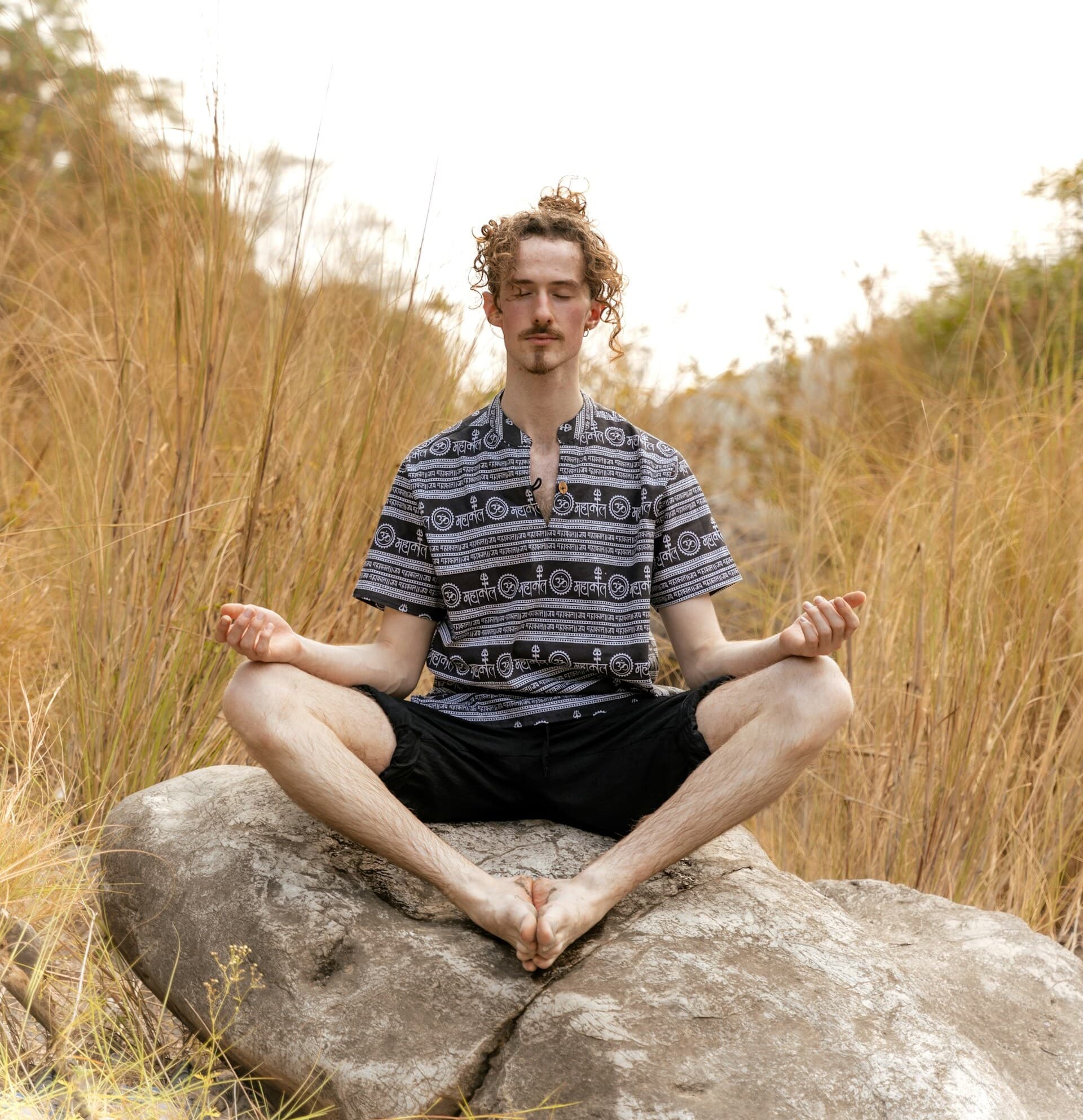 A person sitting cross-legged on a large rock in a peaceful outdoor setting, meditating with eyes closed and hands resting on their knees, surrounded by tall grass and natural serenity.