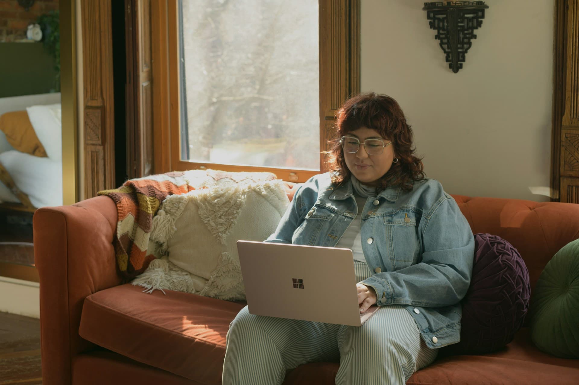 A person sitting on an orange couch with decorative pillows, working on a laptop in a sunlit room. They are wearing a denim jacket and glasses, surrounded by a warm and cozy atmosphere with soft lighting.