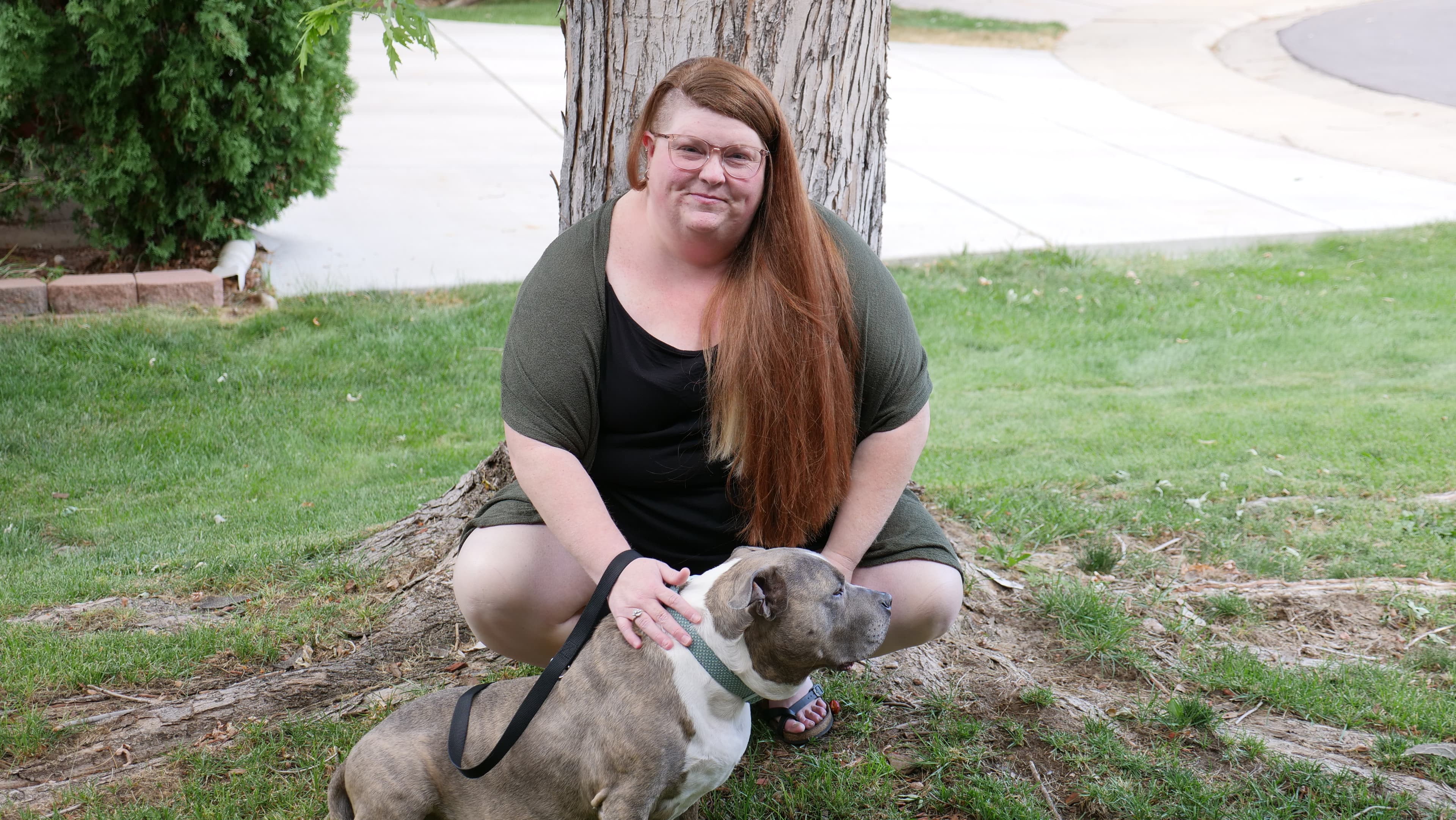 Jamie is kneeling outdoors by a tree with a calm, gray and white dog by her side. She has long, reddish-brown hair and glasses, wearing a casual black outfit with a green cover-up, smiling warmly.