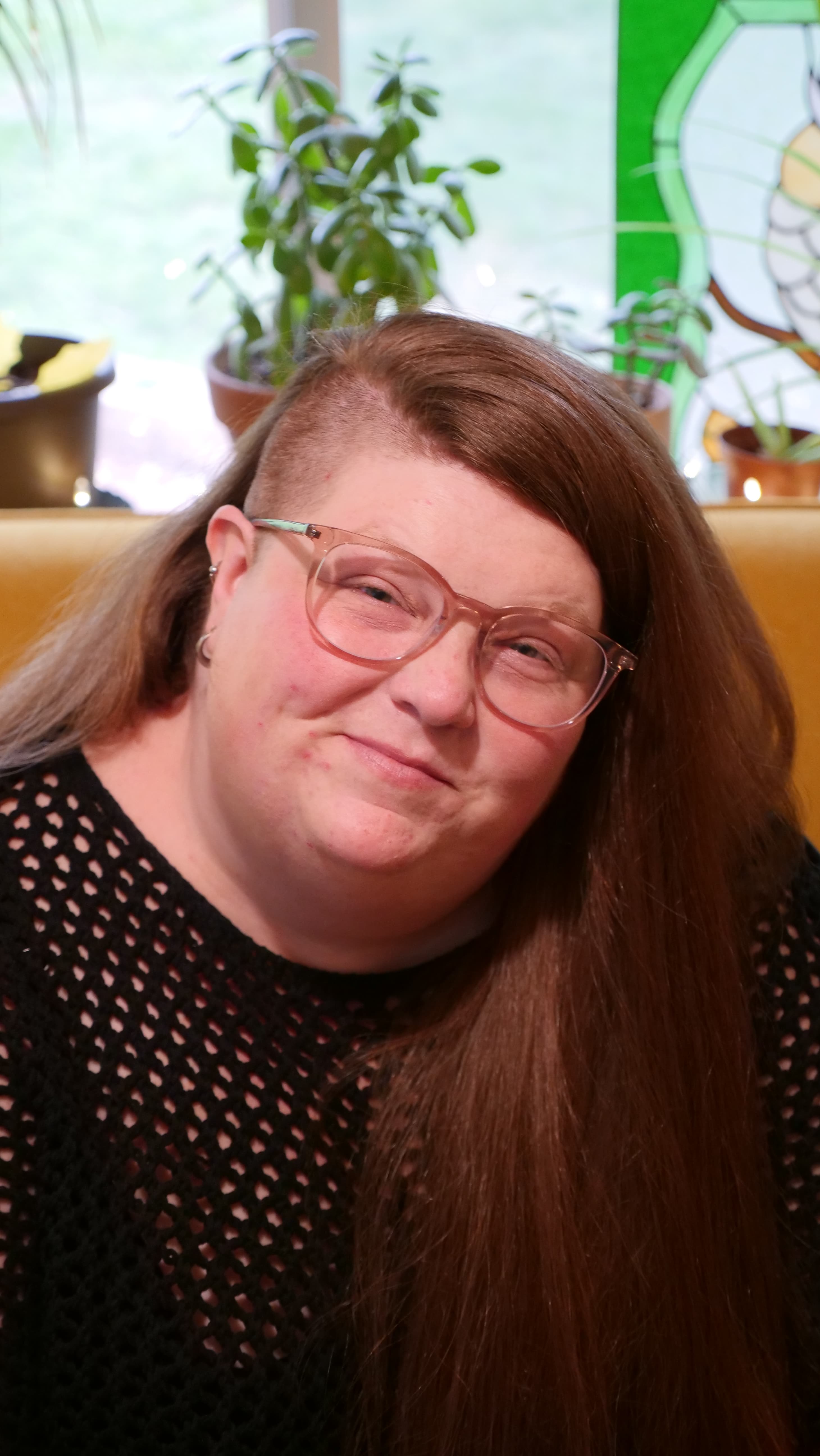 Jamie is smiling warmly while sitting indoors, with long reddish-brown hair and glasses. The background features plants and stained glass, adding to the cozy, welcoming atmosphere.