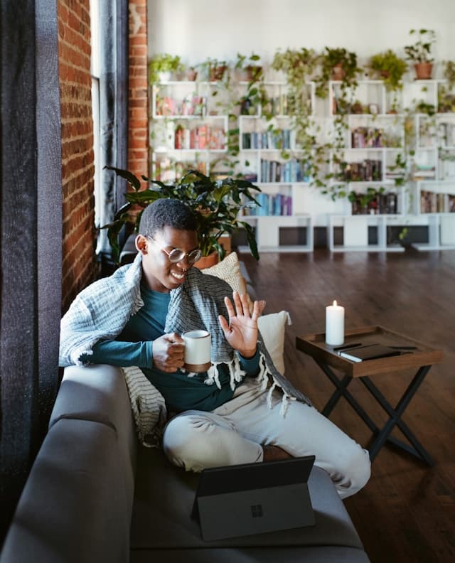 A man sitting on a couch wrapped in a blanket, holding a coffee mug, and smiling while waving at a laptop screen in a cozy, plant-filled room. A lit candle sits on a nearby table, adding to the warm, relaxed atmosphere.