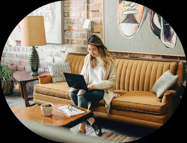 A woman sitting on a vintage-style mustard yellow couch, working on a laptop in a cozy room with exposed brick walls and modern art. A lamp and coffee table with a plant and magazine complete the warm, stylish setting.