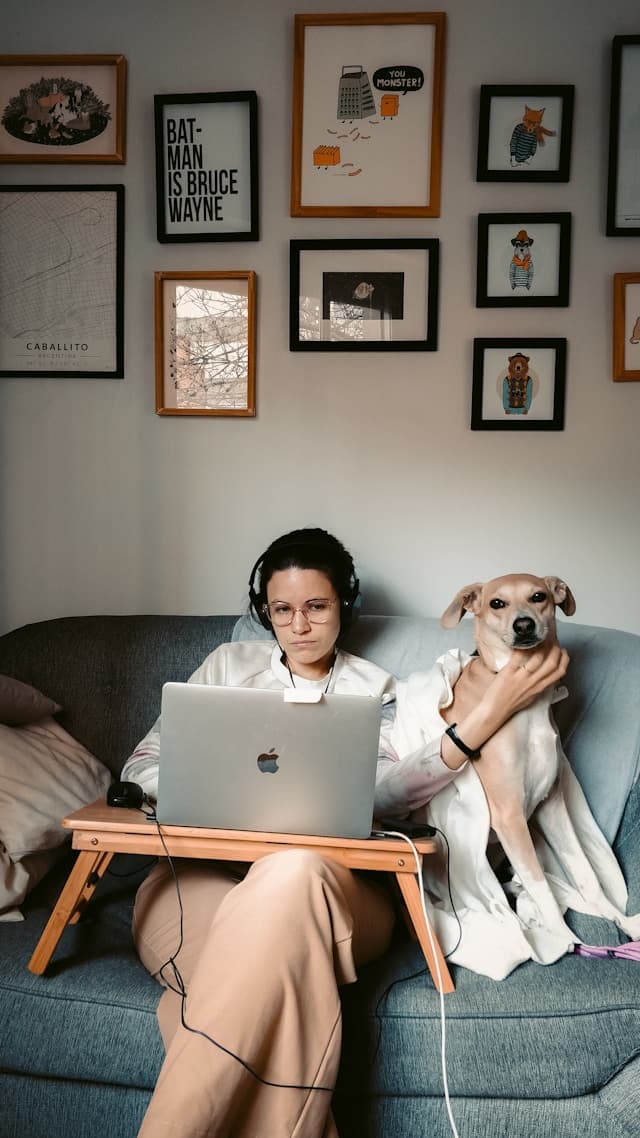 A woman sitting on a couch with a laptop on her lap, wearing headphones, and holding her dog close. The wall behind her is decorated with various framed artwork, creating a cozy and personal atmosphere.