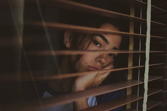 A young woman rests her head on her hand while gazing through window blinds. The dim lighting and her contemplative expression evoke a sense of introspection or quiet reflection.